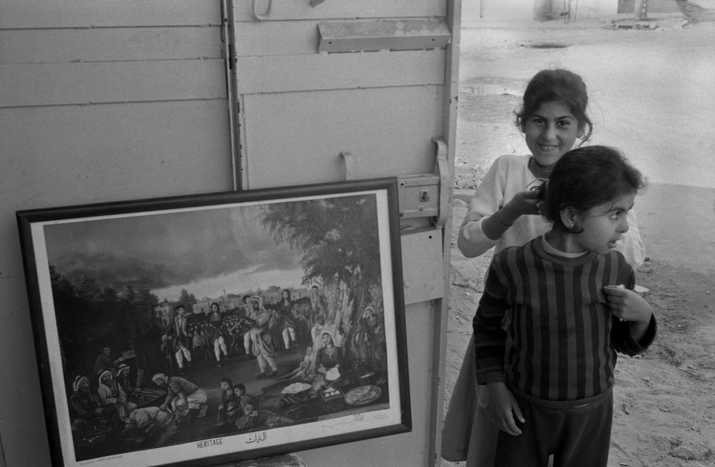 Taken in the Shati Palestinian refugee camp, a glimpse of two girls standing near a shop selling paintings by the Palestinian artist Fathi Ghabin. (1987). From the Joss Dray Collection - The Palestinian Museum Digital Archive.